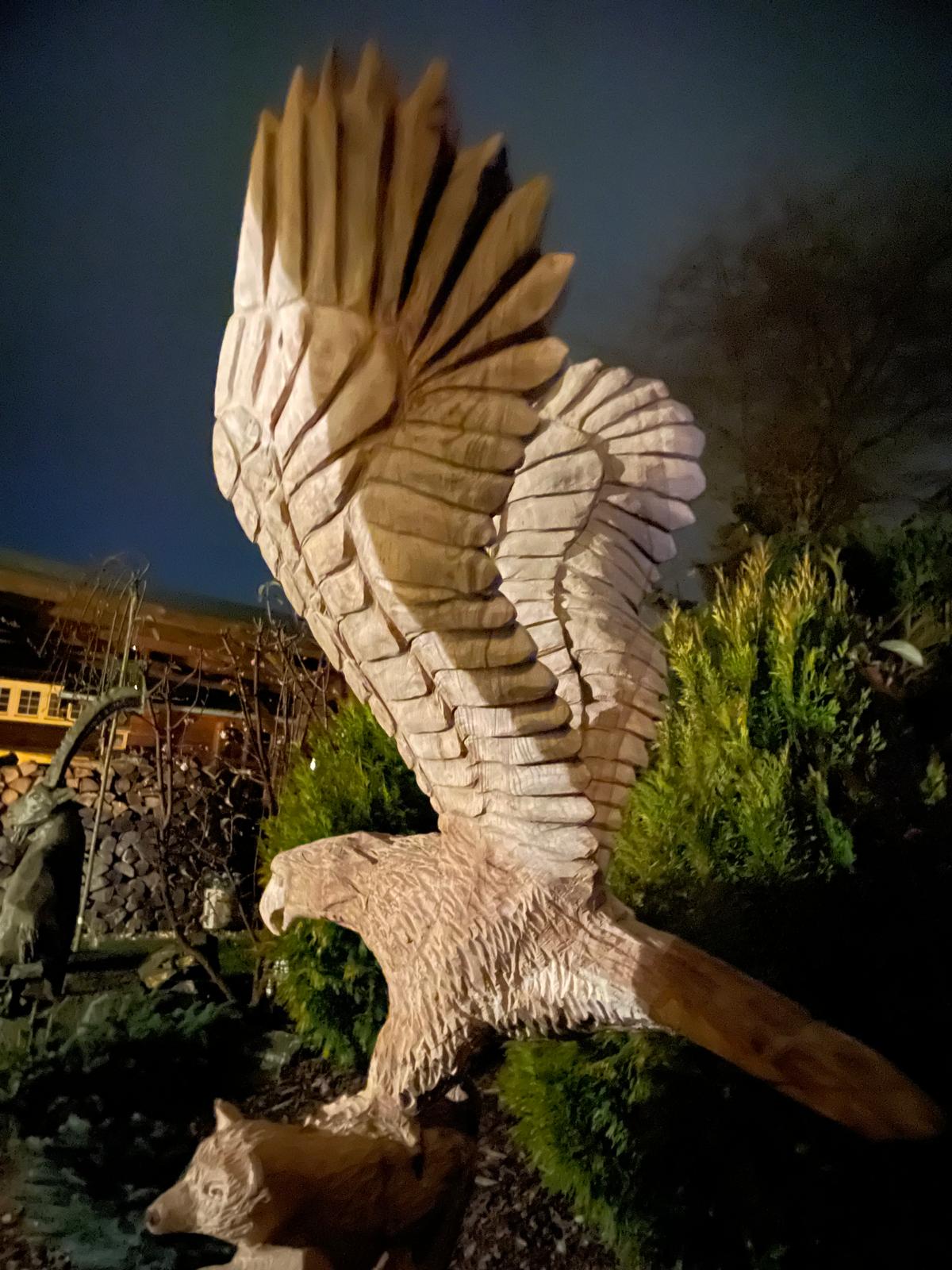 Eine große, detaillierte Kettensägenschnitzerei eines Adlers mit ausgebreiteten Flügeln und einem Fuchs im Außenbereich.